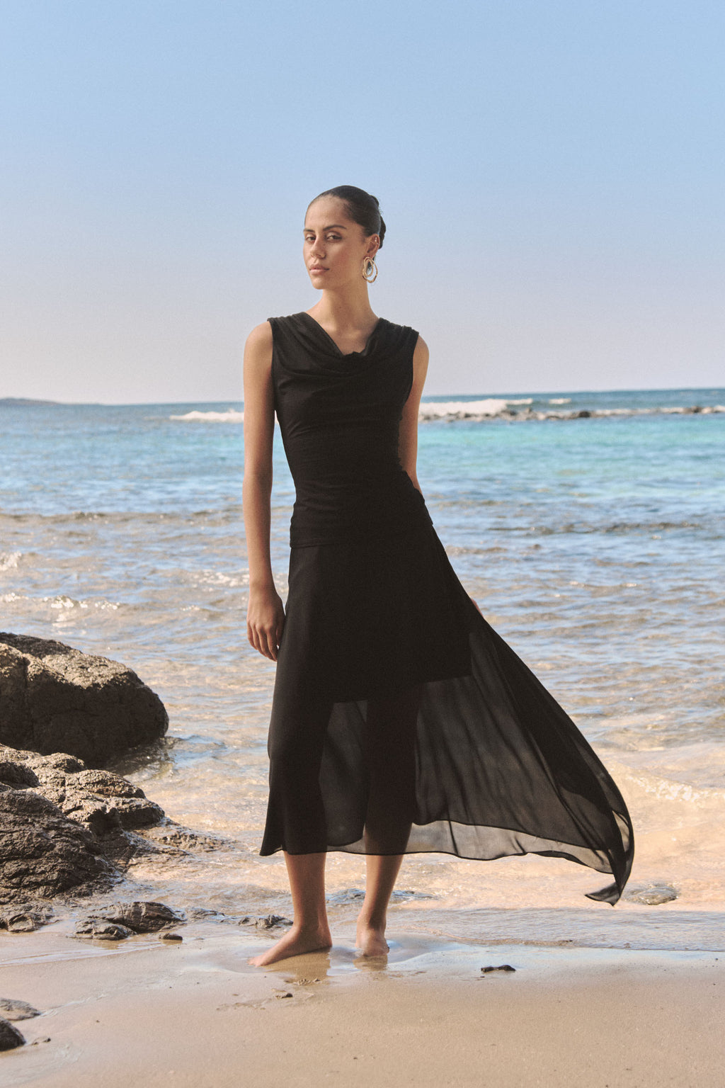 Desiree Asymmetric Skirt in black worn on the beach with a black sleeveless top, skirt blowing in the wind